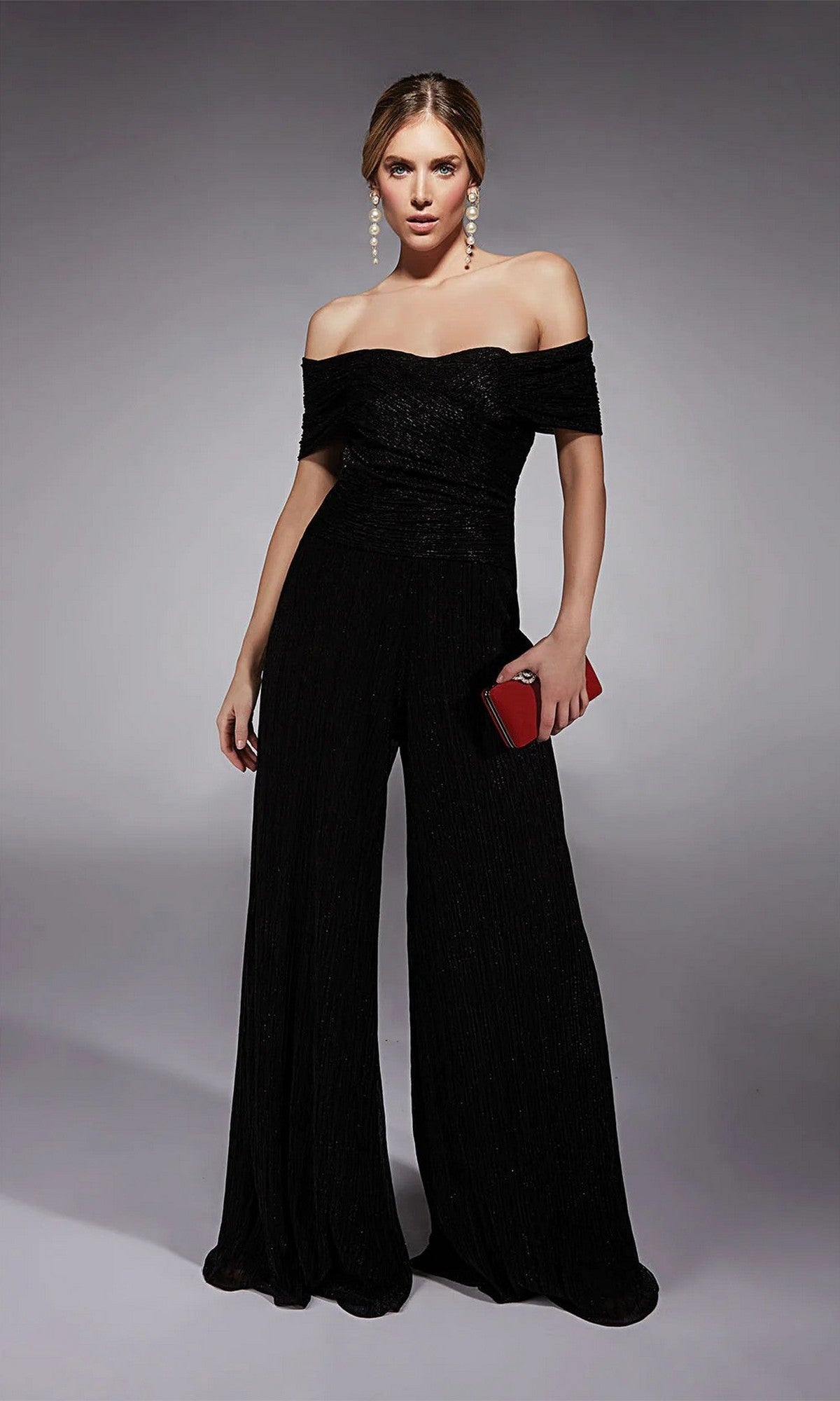 A woman stands against a neutral background, holding a red clutch and wearing the Alyce 27768 Long Formal Jumpsuit—an elegant off-the-shoulder black design perfect for special occasions.